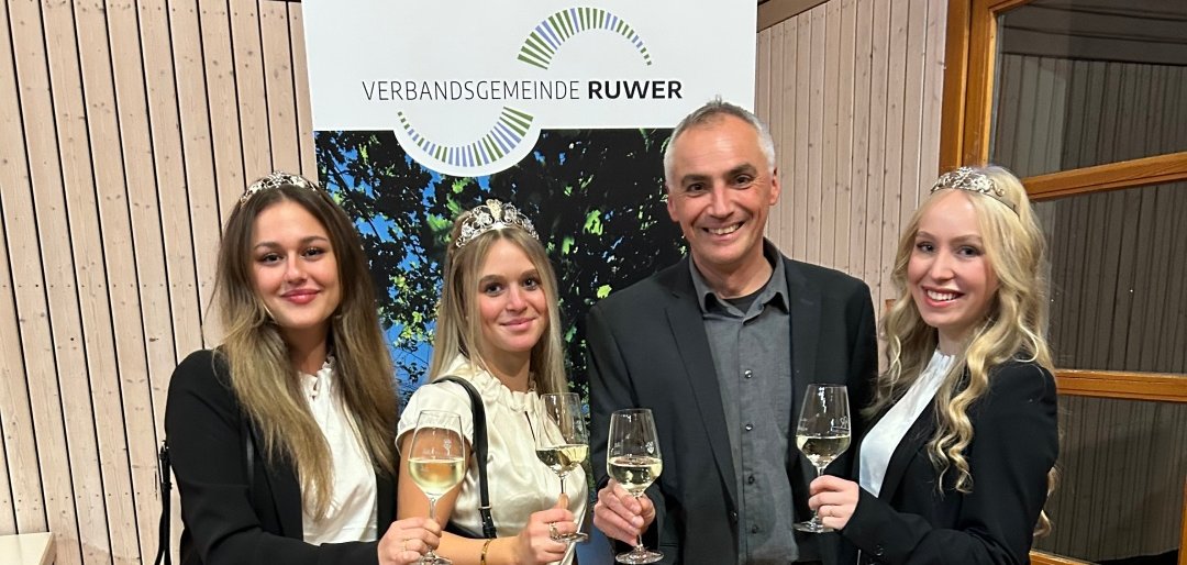 4 Personen, 3 Frauen, 1 Mann lächeln in die Kamera und haben jeweils ein Glas Wein in der Hand. Im Hintergrund steht ein Roll-Up mit dem Logo der VG Ruwer. Die Personen sind der zukünftige Bürgermeister Thomas Hoffmann und die Ruwer-Weinmajestäten.