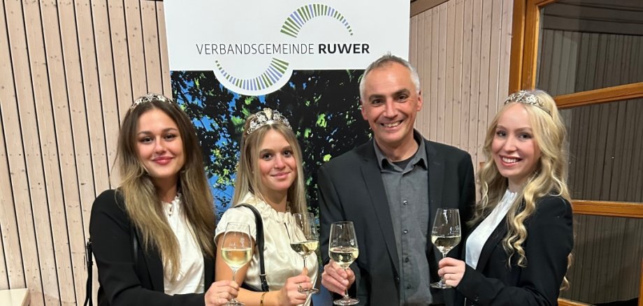 4 Personen, 3 Frauen, 1 Mann lächeln in die Kamera und haben jeweils ein Glas Wein in der Hand. Im Hintergrund steht ein Roll-Up mit dem Logo der VG Ruwer. Die Personen sind der zukünftige Bürgermeister Thomas Hoffmann und die Ruwer-Weinmajestäten.