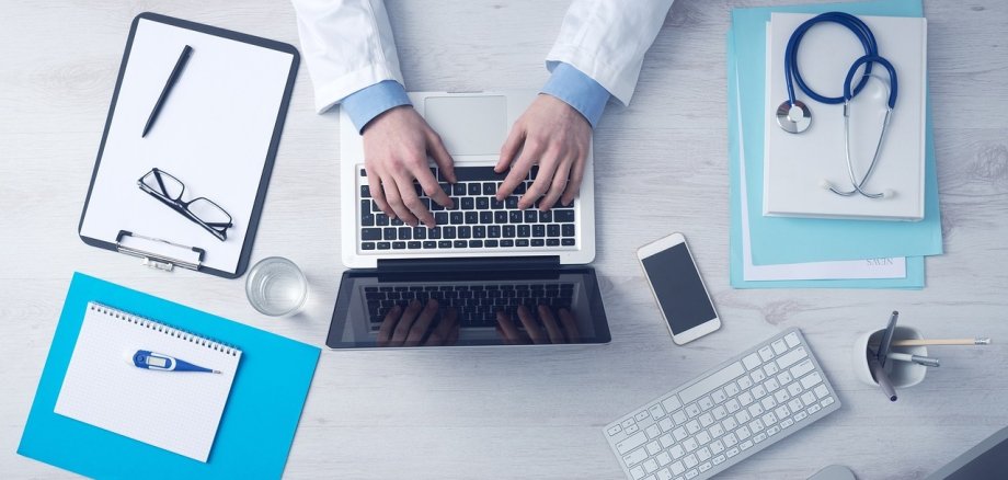 Arzt tippt Diagnose auf Laptop, weitere Utensilien aus der Arztpraxis.