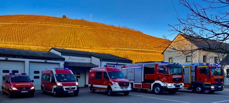 Feuerwehrhaus mit mehreren roten Einsatzfahrzeugen davor, im Hintergrund ein Hügel mit herbstlicher Vegetation.