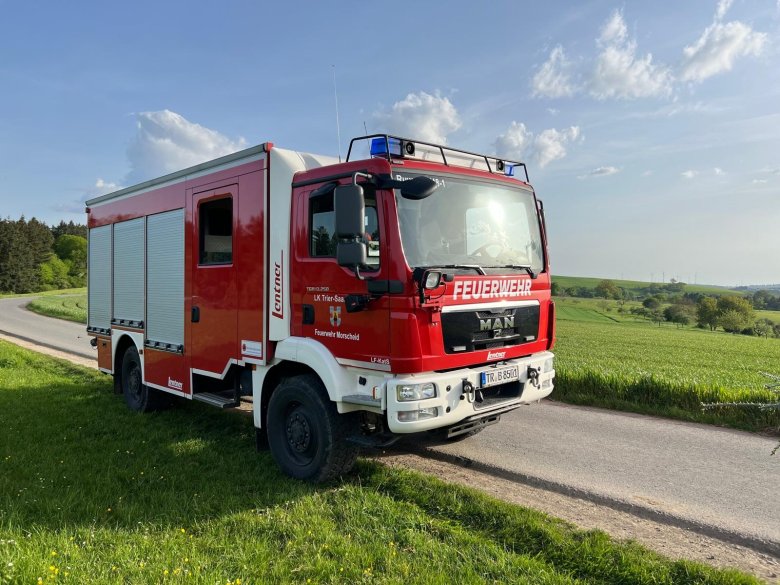 Rotes Löschfahrzeug der Feuerwehr auf einer Landstraße vor ländlicher Landschaft.